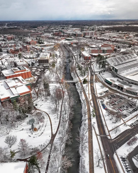 Aerial view of the Red Cedar River winding through Michigan State University\u2019s snow-covered campus, with Spartan Stadium visible on the right.
