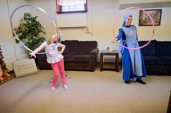 A Religious Sister of the Daughters of Mary of Nazareth is standing in a room with a young girl. Each one is twirling a large hula hoop around her right arm and smiling at the camera. The young girl is on the left side of the picture and wearing pink slacks with a white rainbow ruffled shirt. The Sister is on the right side of the picture and wearing a dark blue and light blue habit with light blue veil, white rope belt, and black shoes.