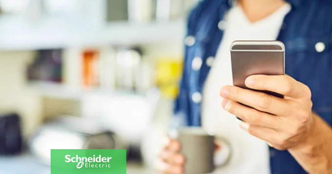 Nahaufnahme einer Person in einem Jeanshemd, die in der rechten Hand ein Smartphone und in der linken Hand einen grauen Becher h\u00E4lt, vor einem unscharfen Hintergrund.
