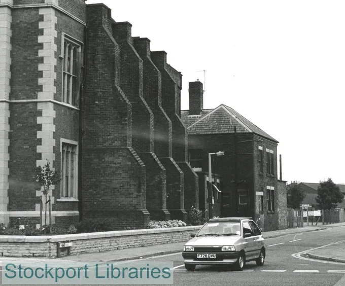 A silver car drives is at a T junction, with to the left a brick building with stepped buttresses.