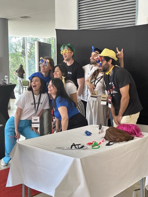 Varias personas asistentes y colaboradoras de Commit Conf posan juntas en un fotomatón durante el evento. Llevan accesorios divertidos como gafas de colores, sombreros y pelucas mientras sonríen frente a la cámara. En primer plano hay una mesa con más complementos para las fotografías y, al fondo, un panel negro utilizado como fondo para las imágenes. La escena transmite un ambiente distendido, cercano y festivo.