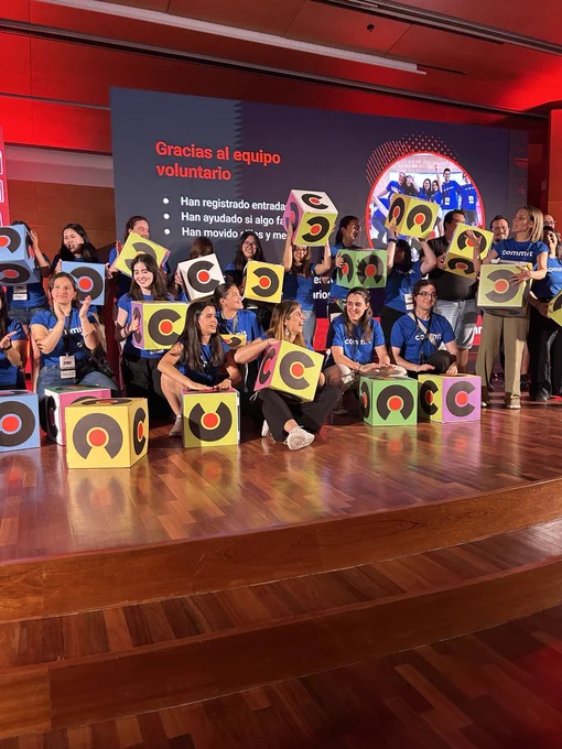 Fotografía del equipo de voluntariado de Commit Conf sobre el escenario durante el cierre del evento. Varias personas vestidas con camisetas azules de la organización posan sosteniendo cubos decorados con el logotipo de la conferencia. Al fondo se proyecta una pantalla con un mensaje de agradecimiento al voluntariado. La imagen transmite celebración, compañerismo y reconocimiento al trabajo realizado por quienes han hecho posible el evento.