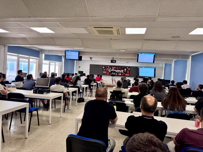 Aula llena de asistentes durante la sesión de Adrián Ferrera en  Commit Conf. Las personas participantes están sentadas frente a un ponente que expone contenido técnico apoyándose en varias pantallas de presentación. La sala cuenta con mesas equipadas con ordenadores y un gran cartel de Commit Conf al fondo. La imagen refleja un momento de aprendizaje, intercambio de conocimiento y participación activa dentro del evento.