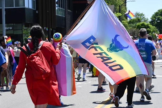 A person in a red jacket & backpack walks in a sunny parade, holding a large rainbow flag featuring 'BUFFALO' and a bull logo. Other participants, city buildings, & a distant clock tower are visible. Yellow road lines mark the pavement.