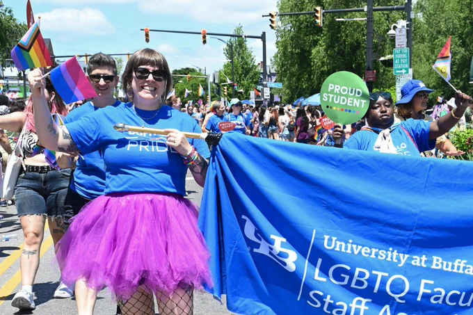 A smiling person in a blue \"PROUD\" shirt, purple tutu, and fishnets holds a pole for a blue banner reading \"University at Buffalo LGBTQ Faculty Staff Association.\" They also hold pride flags in a sunny parade, while another person carries a \"PROUD STUDENT\" sign.