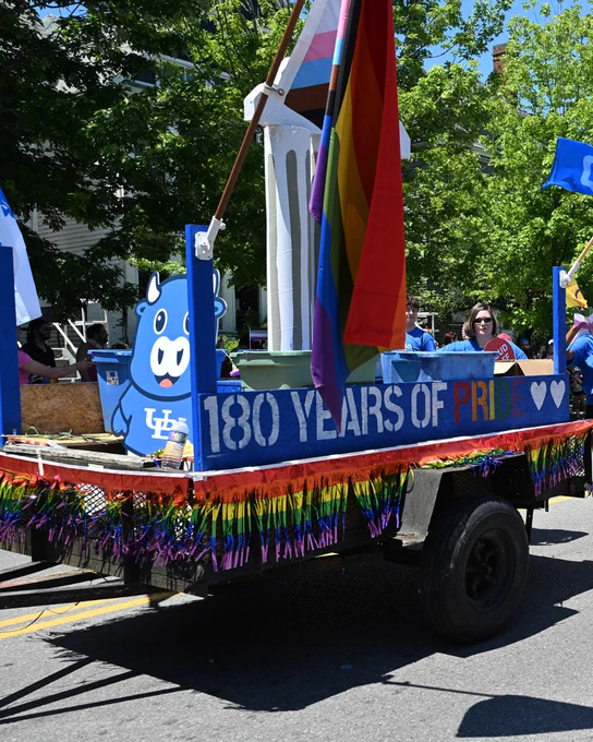 A blue parade float with '180 YEARS OF PRIDE' painted on its side, decorated with rainbow fringe. A blue cartoon bull mascot is on the front. Large pride flags wave from the float. People in blue shirts are on board, one holding a 'PROUD' sign.