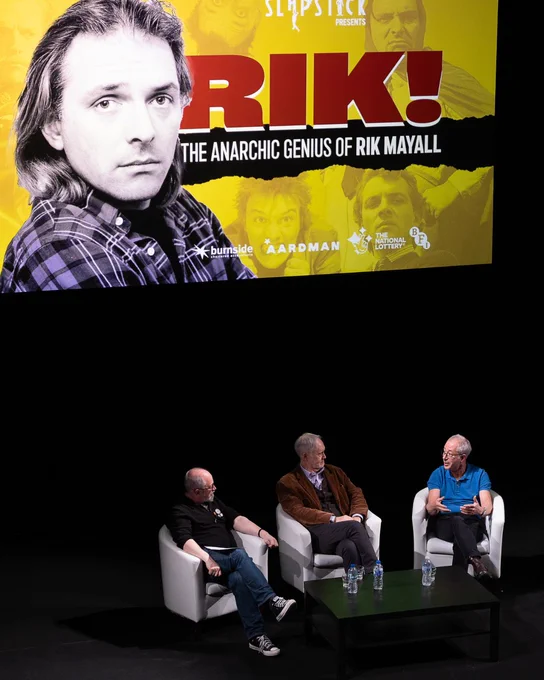 A picture of Robin Ince, Nigel Planer, and Ben Elton in conversation on stage, with a poster of the event featuring Rik Mayall