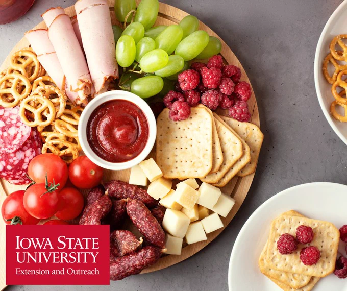 Round wooden board with rolled turkey slices, green grapes, raspberries, crackers, cheese cubes, cherry tomatoes, salami, pretzels, and a bowl of red dipping sauce.