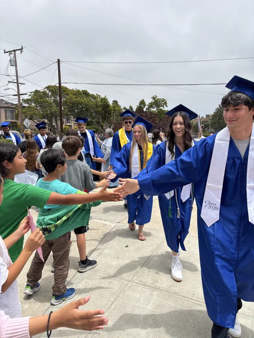 Graduating seniors in blue caps and gowns participate in a school walk, exchanging high-fives with younger students and families gathered outside.