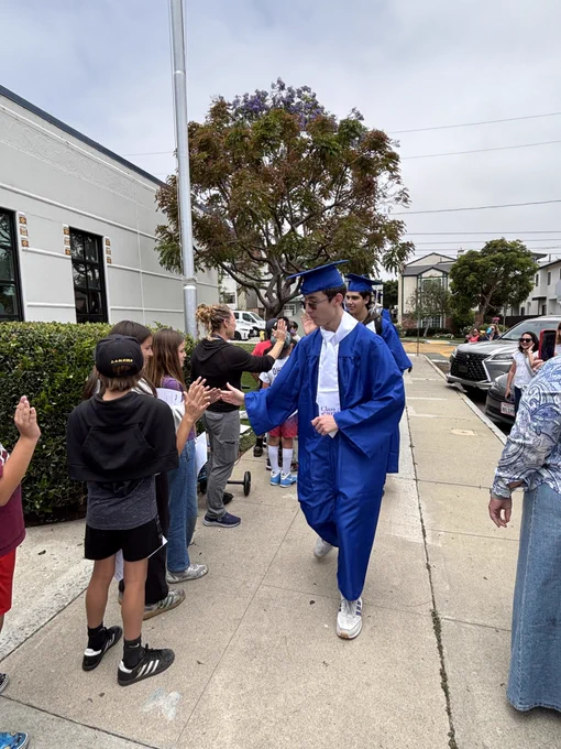 Graduating seniors in blue caps and gowns participate in a school walk, exchanging high-fives with younger students and families gathered outside.