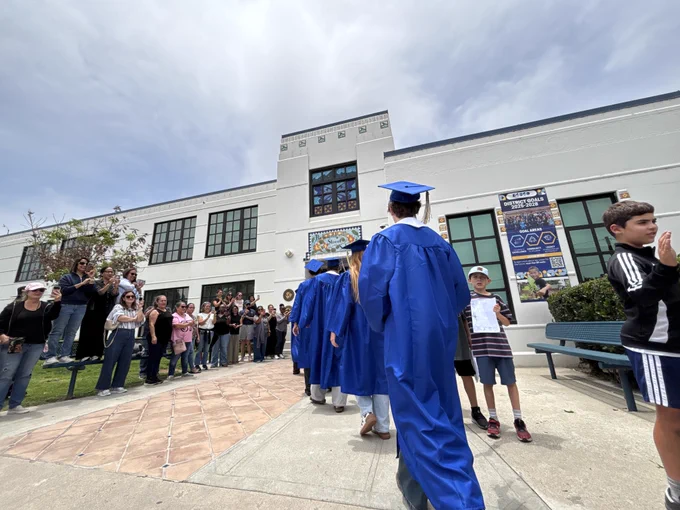 Graduating seniors in blue caps and gowns participate in a school walk, exchanging high-fives with younger students and families gathered outside.
