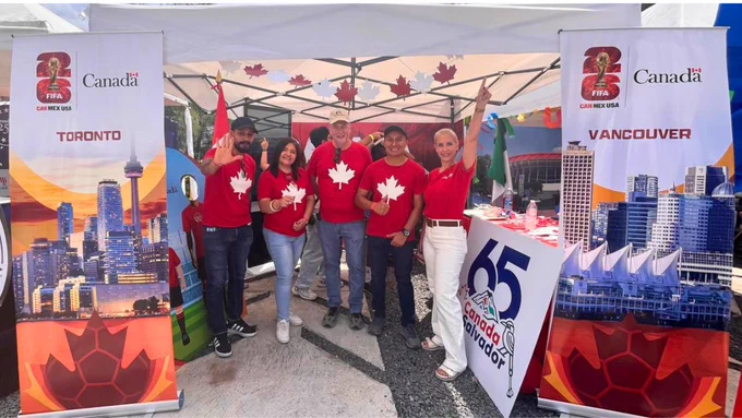 Five smiling people standing in front of the Embassy of Canada booth at a soccer fair. They are wearing red shirts with white maple leaves. Banners highlight Toronto and Vancouver as FIFA World Cup 2026 host cities.