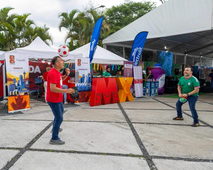 Two men playfully heading a soccer ball at an outdoor festival. One wears a red Canada shirt, the other a green Mexico jersey. The Canada booth and a large "MX" sign are visible in the background.