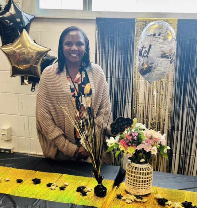 A woman smiles at a decorated table with flowers and balloons, suggesting a celebratory atmosphere. The setting appears welcoming and joyful.