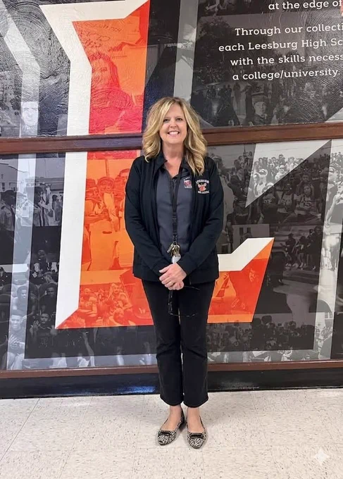 A woman stands smiling in front of a mural at Leesburg High School. The background consists of a large 'L' with text and monochrome photos of students.