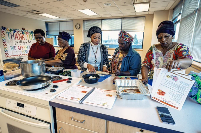 Five women in African attire and headress stadning at a counter in a kitchen reviewing some meal planning.