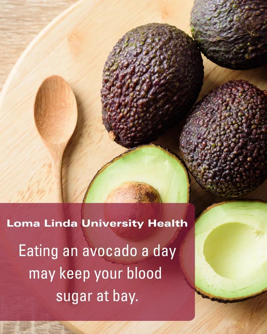 A wooden table with a spoon, whole avocados, and a halved avocado. Text overlay reads: "Loma Linda University Health: Eating an avocado a day may keep your blood sugar at bay."