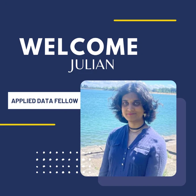 Welcome Julian. Applied Data Fellow. Julian has short black hair, is wearing a blue button up blouse, necklaces, and triangle shaped earrings. In the background is a body of water and trees.