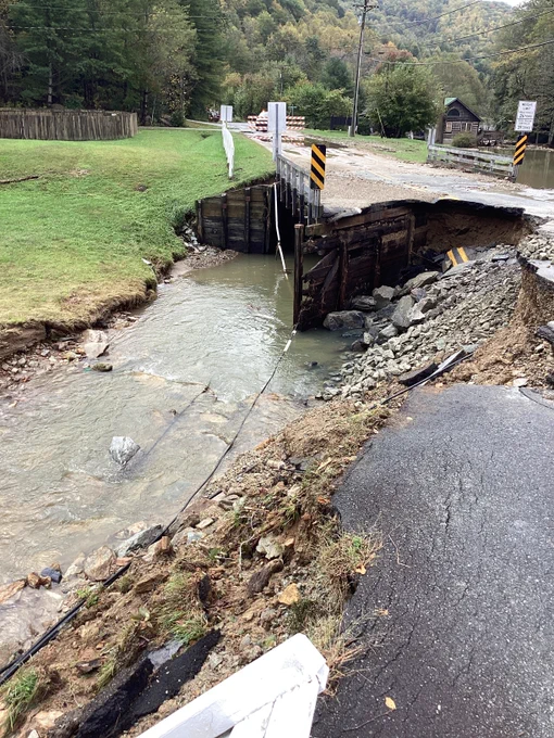 A damaged road with a section collapsed into a flowing stream below. Traffic signs and barriers are placed around the damaged area for safety. Trees and a small building are visible in the background.