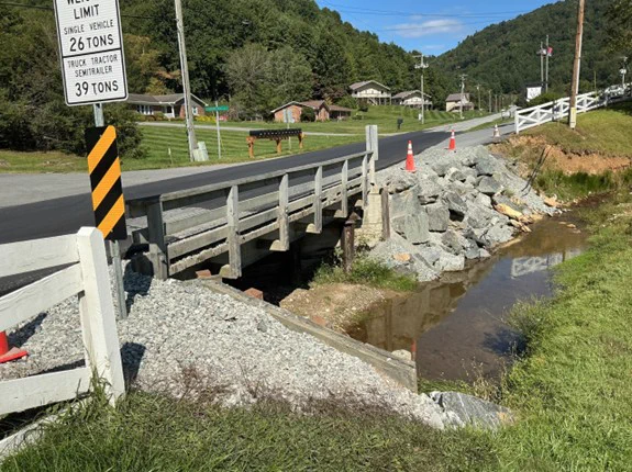 A small bridge with a load limit sign crosses a narrow creek alongside a rural road. Traffic cones and large rocks are near the bridge. In the background, there's a grassy area with houses and a forested hillside.