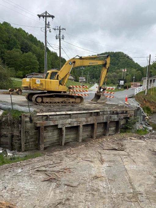 A yellow excavator is parked on a partially collapsed bridge, surrounded by orange and white road barriers. There are orange traffic cones nearby. In the background, utility poles and green, tree-covered hills are visible under a cloudy sky.