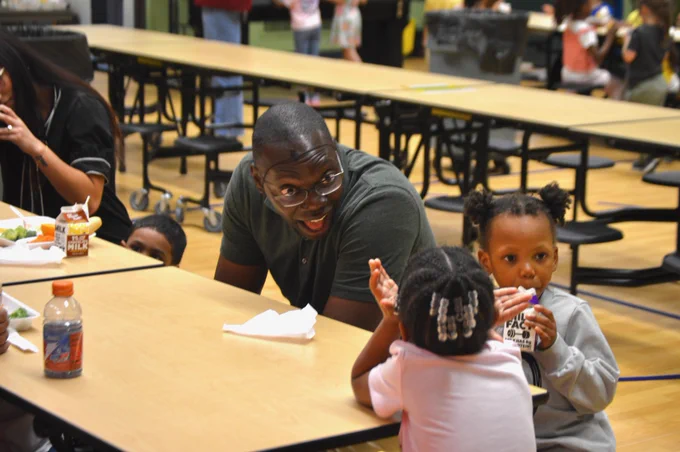 The Lieutenant Governor, wearing glasses and a dark green polo shirt, leans across a wooden school cafeteria table to cheerfully talk with young children. He has a wide, happy expression as he interacts with a young girl with braided hair and another student holding a small milk carton. The background shows an open school cafeteria with long tables and stools.