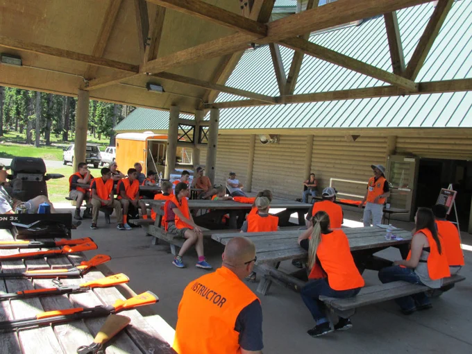 A group of hunter education students, all wearing orange high vis vests, sitting on outdoor picnic tables listening to a hunter education instructor address the class.