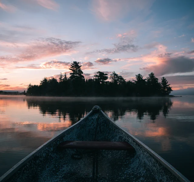 Photo of the bow of a weathered canoe heading toward a small island covered with fir trees, on a calm lake at either sunrise or sunset.  The sky is pale blue, with some clouds that are lavender, peach, pink, and dark gray.