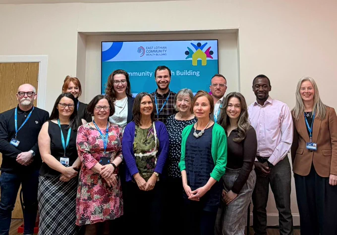 A diverse group of thirteen people smiling for a photo in front of a screen displaying "East Lothian Community Wealth Building" logo.