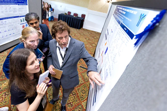 Two men and two women look at a scientific poster. One of the men is pointing to a spot on the poster and talking about the data that is shown while one of the women takes notes.