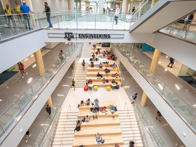 A spacious, multi-level interior view of the Texas A&M Engineering building. People are seated and walking in the open atrium area with wide, tiered wooden steps. Glass railings line the upper walkways, and the space is well-lit with natural light.