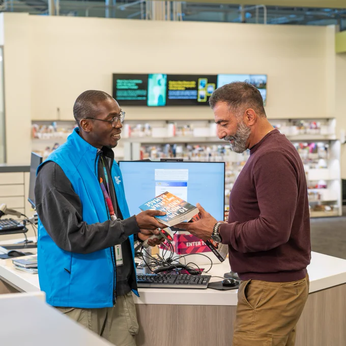 A person wearing a blue vest is assisting another individual with books at a library counter. They are holding a book and discussing it. The background features bookshelves and a digital display screen.