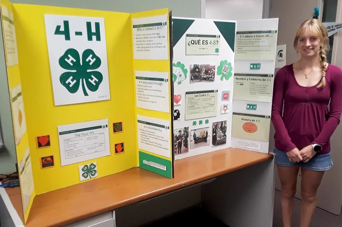 A student stands beside a tri-fold educational display about 4-H featuring bilingual English and Spanish information, photos, and the organization’s four-leaf clover emblem. The presentation board includes sections about 4-H history, membership, and programming, displayed on a table indoors.