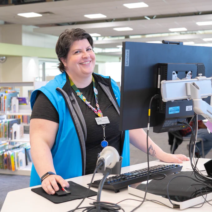 Person wearing a blue vest stands at a check out desk at a Library. 