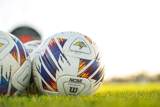 Close-up of two NCAA soccer balls resting on green grass, featuring multicolored patterned panels and a South Dakota State jackrabbit logo.