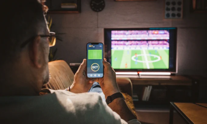 A person is sitting on a couch watching a soccer match on a TV screen. The person is holding a smartphone displaying a betting app interface with a "BET" button. 