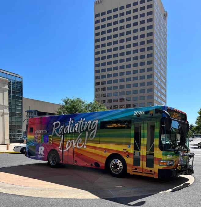 City bus painted with a vibrant rainbow design and the words 'Radiating Love!' parked near tall office buildings on a sunny day.