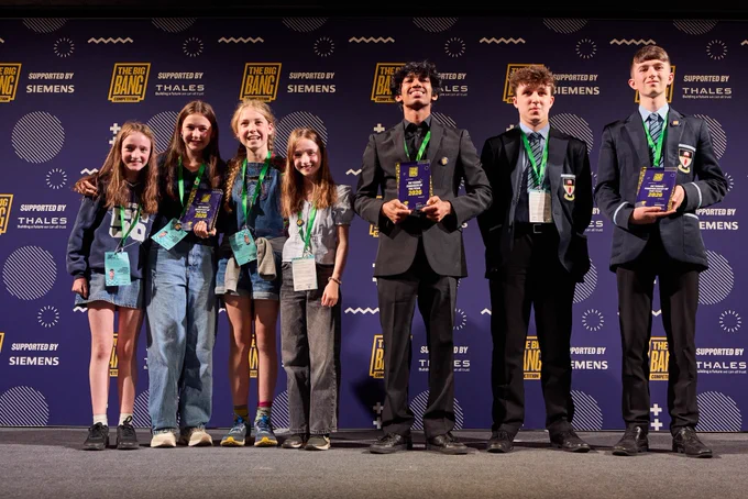 A group of young individuals stand on a stage, holding awards at The Big Bang event, supported by Siemens and Thales. They are smiling in front of a purple backdrop with event logos. They wear lanyards and formal attire.