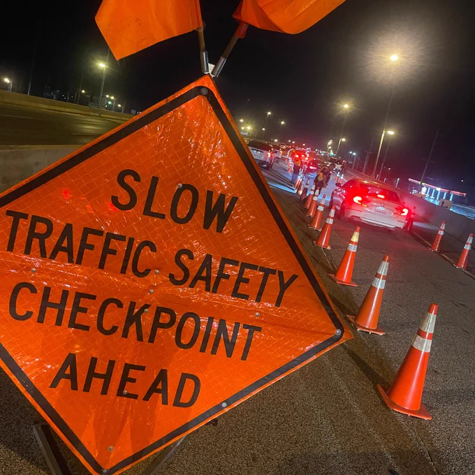 A nighttime roadway scene shows an orange warning sign in the foreground that reads “SLOW TRAFFIC SAFETY CHECKPOINT AHEAD.” Traffic cones line multiple lanes, guiding a line of vehicles toward a checkpoint area illuminated by streetlights. Several people are visible near the cones as cars move slowly through the controlled traffic pattern.