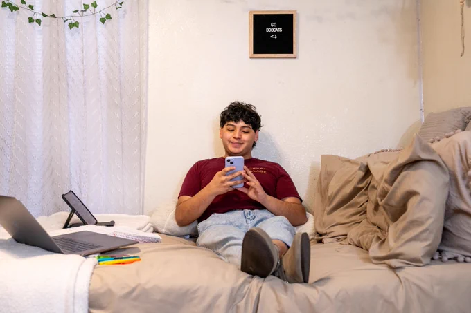 Person relaxing on a bed using a smartphone, surrounded by pillows and a laptop on the side table in a cozy room.