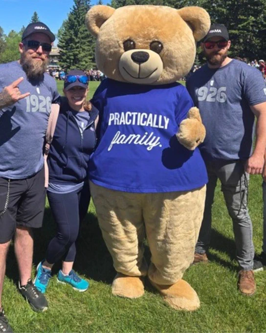 Large teddy bear mascot wearing a blue shirt with 'Practically family.' printed, standing outdoors on grass with three people in casual clothing around it.