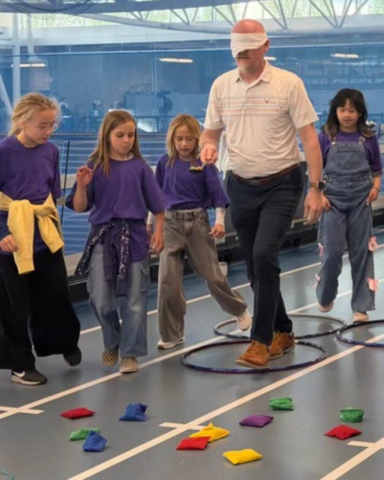 A group of children and an adult participate in an indoor activity involving colorful bean bags and hoops on the floor.