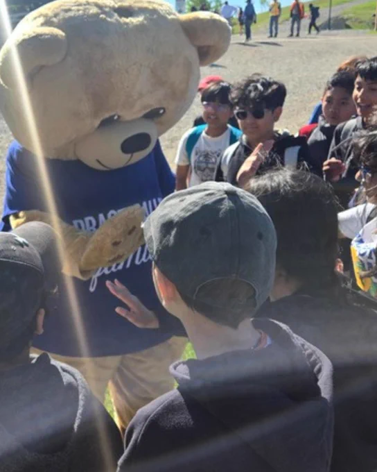 Person in a large teddy bear costume interacting with a group of children outdoors on a sunny day.