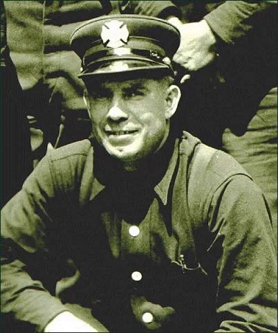 A man in an old-fashioned fireman's uniform sits confidently. He wears a hat with an emblem, and his expression is calm, conveying pride and duty.