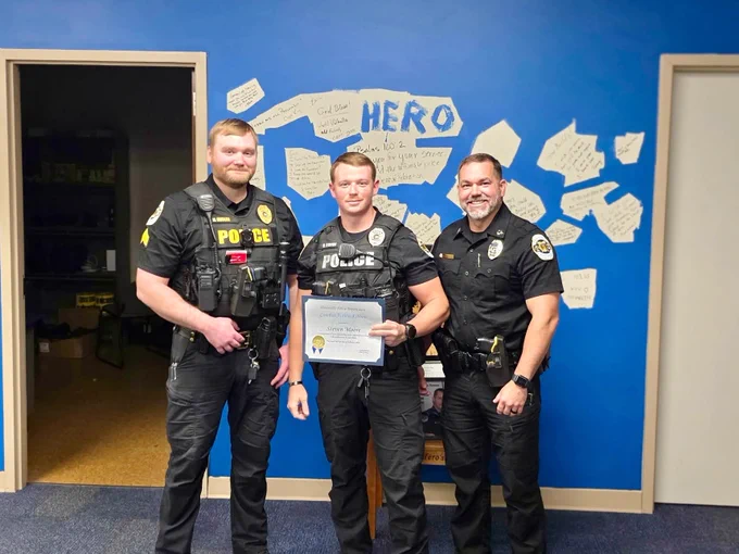 Three individuals in police uniforms are standing together in a room with bright blue walls. The person in the center is holding a certificate with additional text visible below. The wall behind them features a display of handwritten notes arranged around the word 'HERO' in large letters. A doorway to another room is visible on the left.