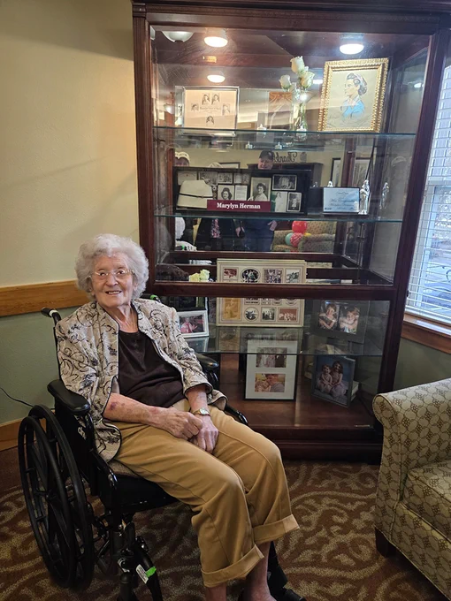 An older woman in a wheelchair smiles as she sits in front of a large cabinet that contains mementos of her life