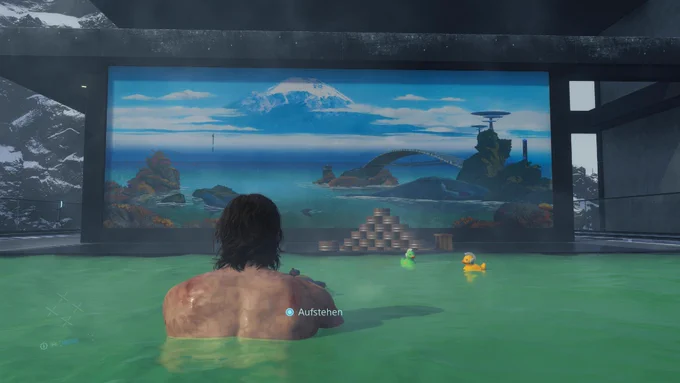 Sam entspannt nach getaner Arbeit in einer heißen Quelle mit grünem Wasser in Death Stranding Directors Cut. Er blickt auf eine virtuelle Berglandschaft während zwei lustige kleine Quietscheentchen neben ihm im Wasser schwimmen.