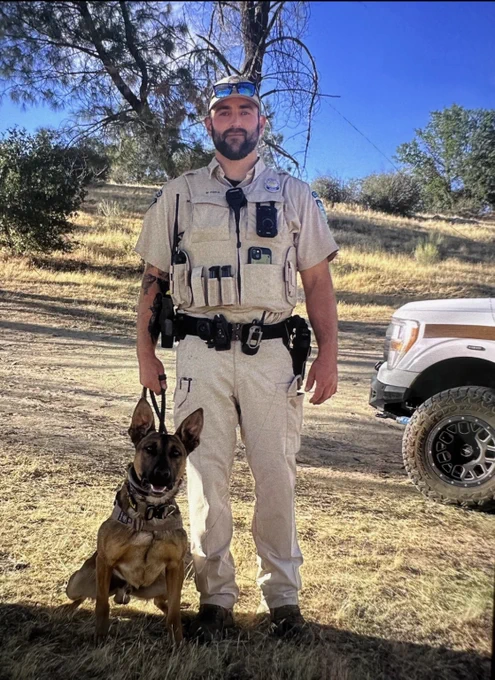 Uniformed officer standing outdoors holding a leash attached to a sitting German Shepherd police dog beside a white vehicle on a sunny day.