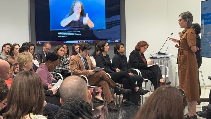 A conference scene with rows of seated attendees facing a speaker at a podium. A panel sits nearby, listening and taking notes. A large screen shows a sign language interpreter against a blue background. People hold papers and look toward the front. 