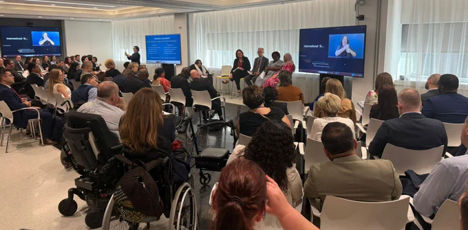A bright conference room filled with rows of seated attendees facing a panel at the front. Several speakers sit on chairs near large screens displaying presentation slides and a sign language interpreter signing. One person stands presenting beside a screen. Some attendees use wheelchairs. The space has white walls, soft lighting, and large windows with sheer curtains, creating a formal, inclusive event setting.